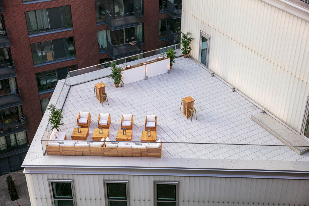 Aerial view of a London rooftop venue featuring modern outdoor seating with wooden furniture, high tables with stools, and decorative potted plants. The terrace, surrounded by residential buildings, offers ample open space and a cozy lounge area for events and gatherings.