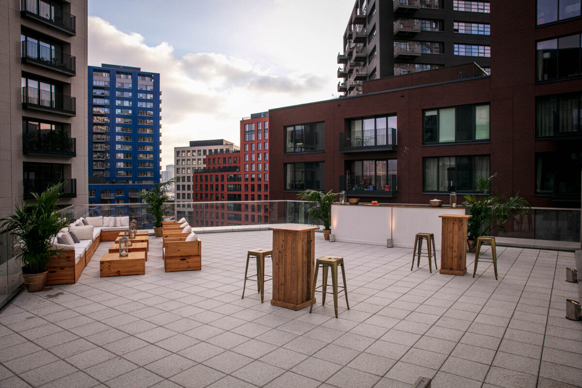 Rooftop venue with modern outdoor seating and tables, surrounded by residential buildings in London. The terrace features a comfortable lounge area with wooden furniture, high tables with stools, and greenery, creating an inviting space for social gatherings and events.