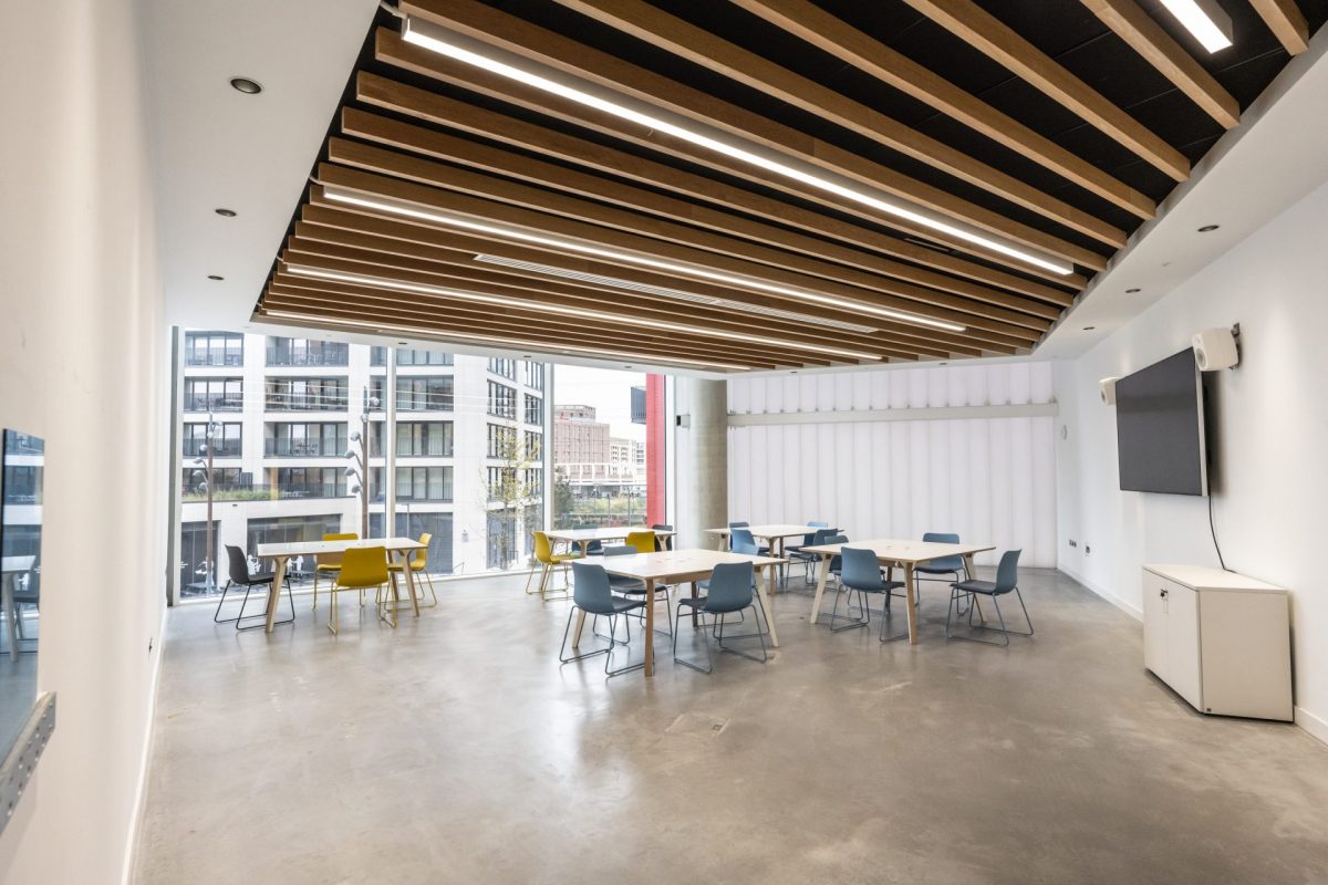 Modern meeting room with large windows, natural light, and a view of city buildings. The room features a stylish ceiling with wooden beams, multiple tables and chairs in a minimalistic setup, and a large wall-mounted screen.