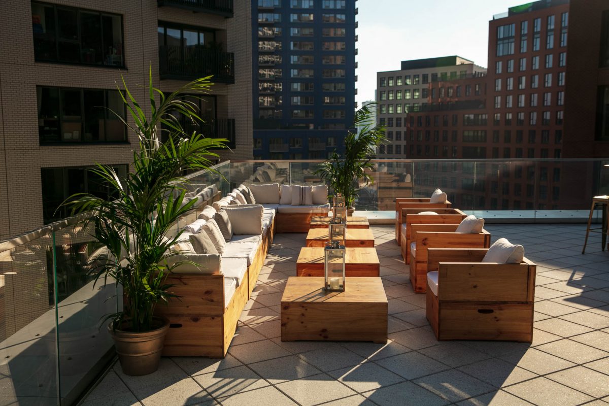 London rooftop venue with cozy wooden lounge seating, potted plants, and lanterns on tables. The space overlooks modern city buildings, offering a serene setting for gatherings and events. The terrace features glass railings and ample seating under natural sunlight.