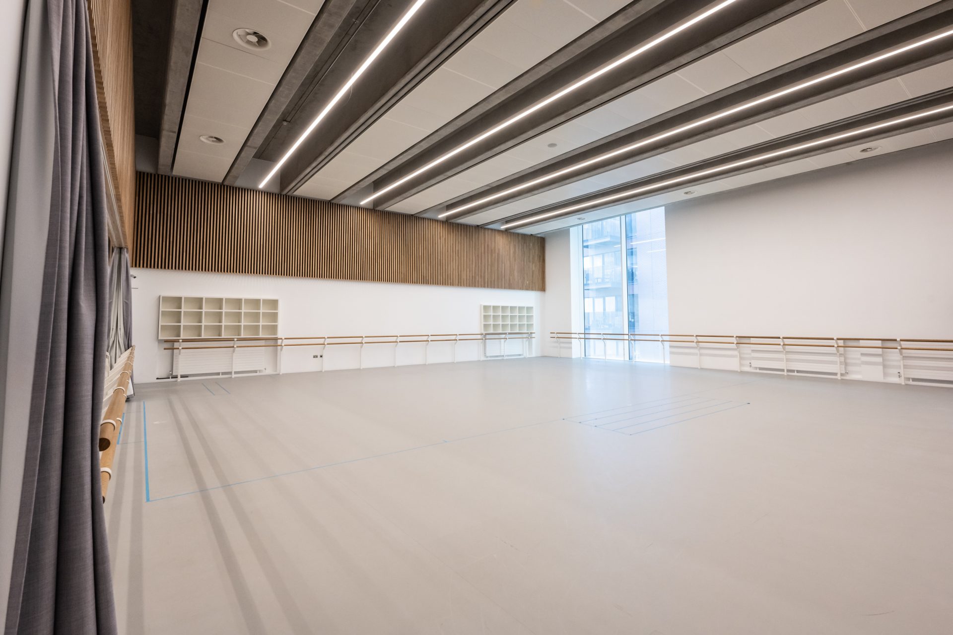 Spacious dance rehearsal studio in London with large windows allowing natural light, wooden barre along the walls, modern lighting, and wooden panel accents on the upper walls.